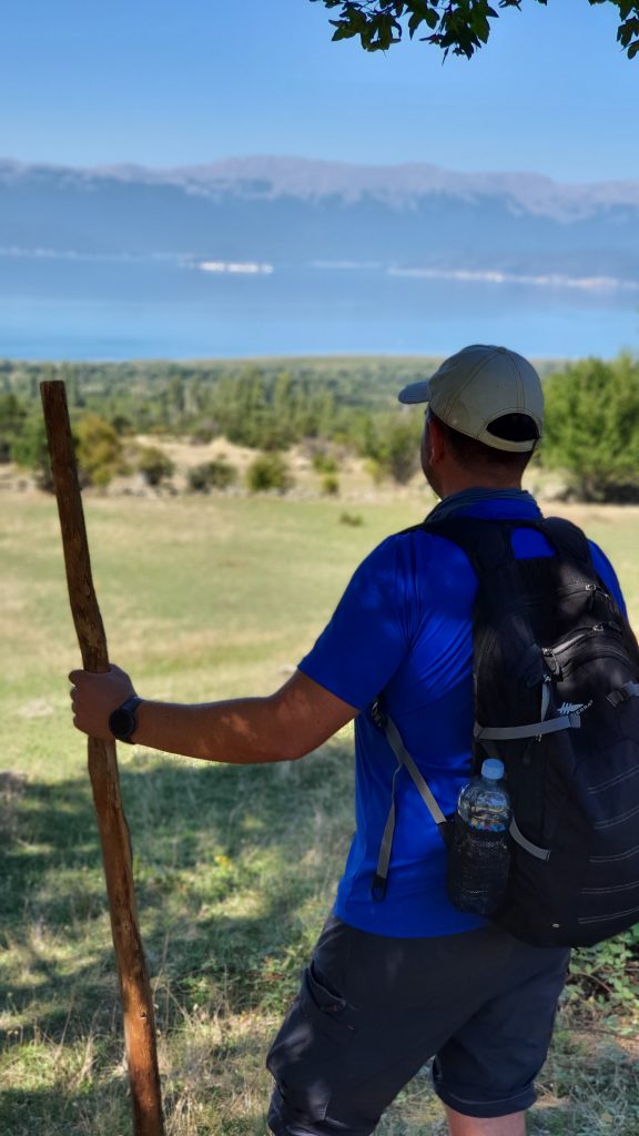hiker with a backpack and wood branch as a stick posing with prespa lake and galicica mountain in a distance